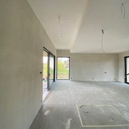 HARMONIA - Remonty i Wykończenia Wnętrz Kamil Łukaszewski - Surowe, jasne wnętrze w trakcie prac: ściany po szpachlowaniu, betonowa posadzka z zaznaczonym obszarem, widoczne przewody elektryczne, duże okna przesuwne z widokiem na zieleń.