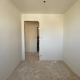 HARMONIA - Remonty i Wykończenia Wnętrz Kamil Łukaszewski - Wnętrze pokoju po szpachlowaniu ścian, widoczne gładkie, białe powierzchnie i niezabezpieczona podłoga z płyty OSB oraz otwór drzwiowy w stanie surowym.