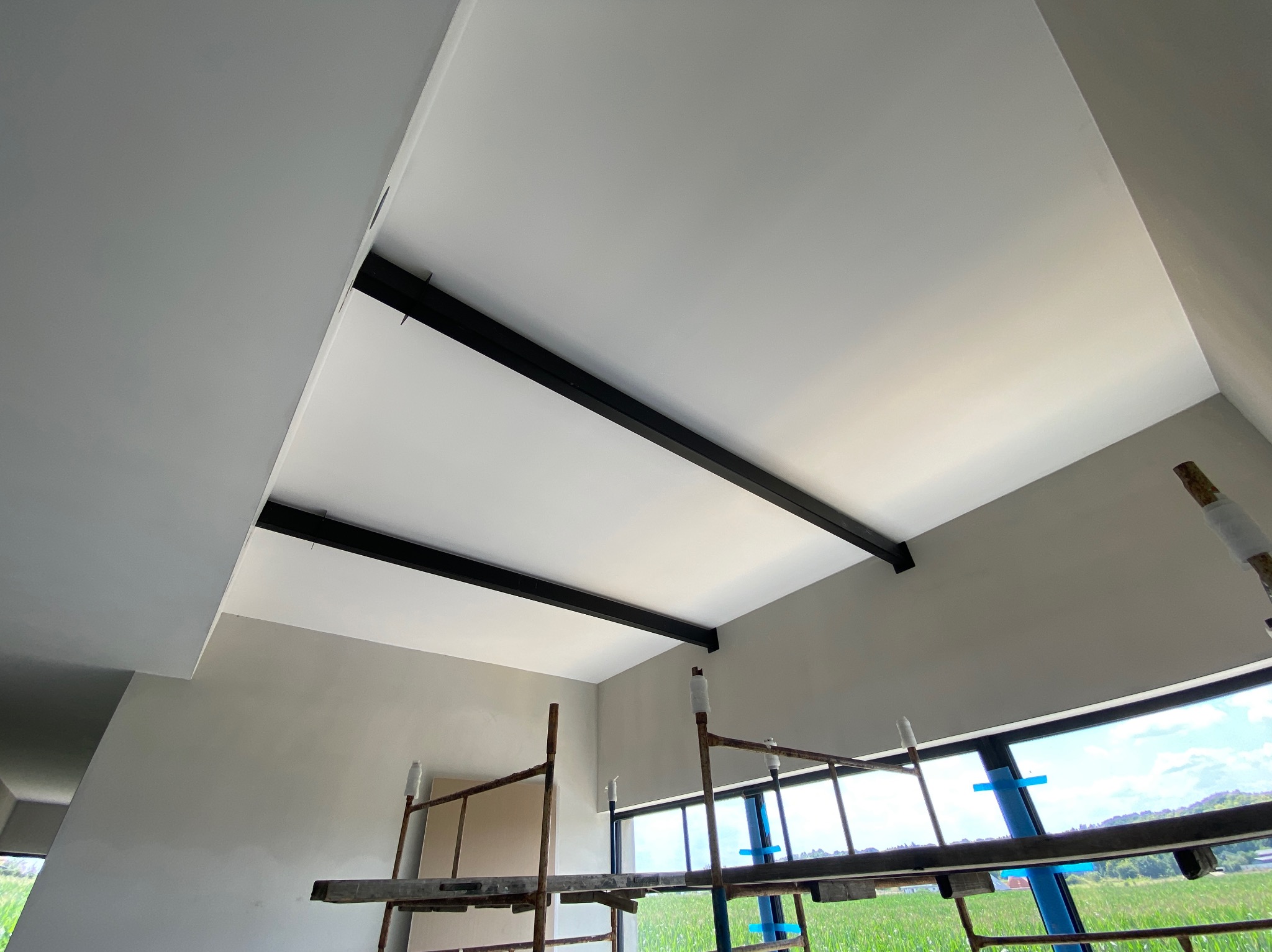 Wnętrze w trakcie prac: pomalowany na biało sufit z czarnymi belkami, szare ściany, widok z dołu na rusztowanie i okno z widokiem na zielone pole.