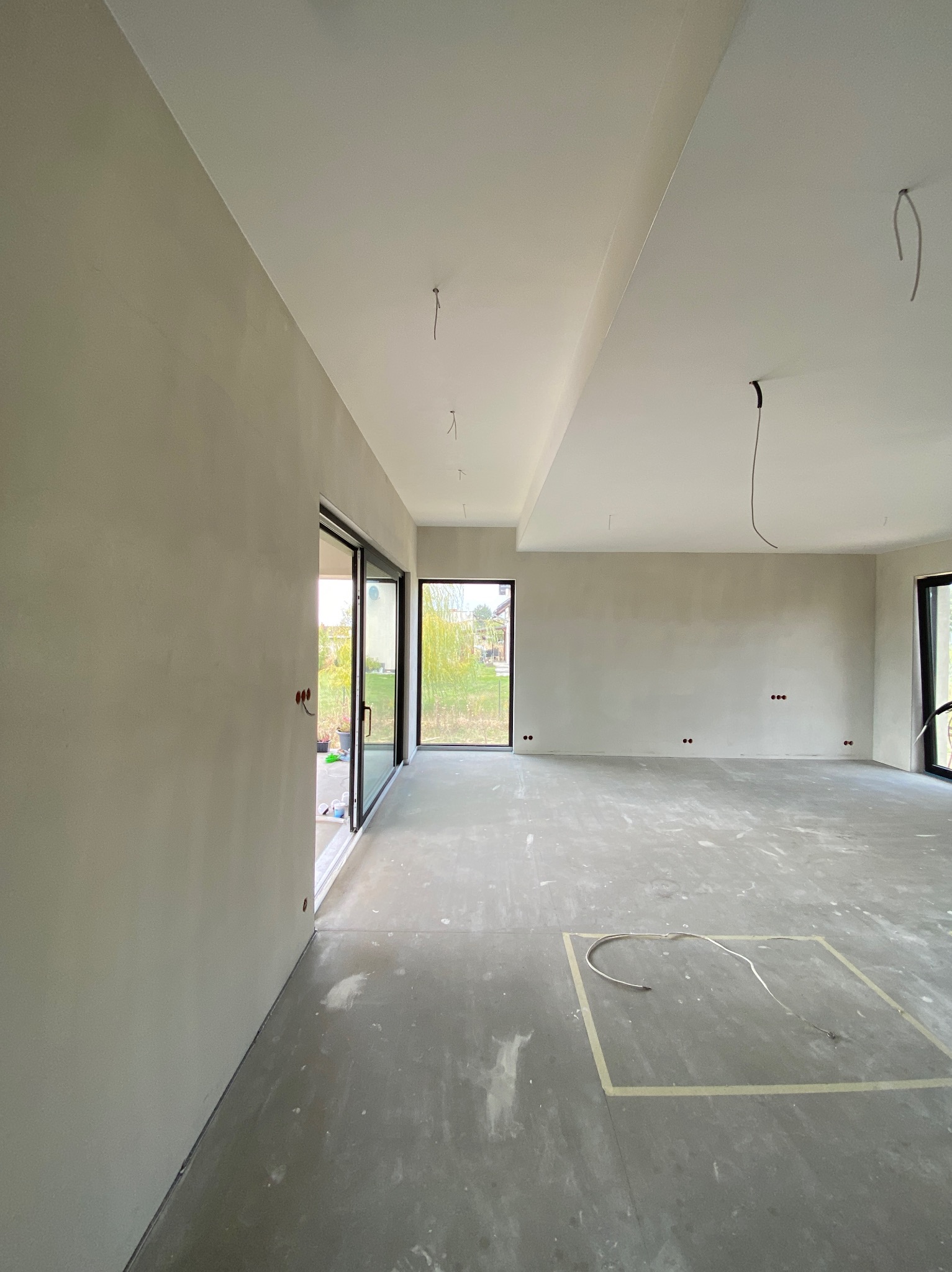 Surowe, jasne wnętrze w trakcie prac: ściany po szpachlowaniu, betonowa posadzka z zaznaczonym obszarem, widoczne przewody elektryczne, duże okna przesuwne z widokiem na zieleń.