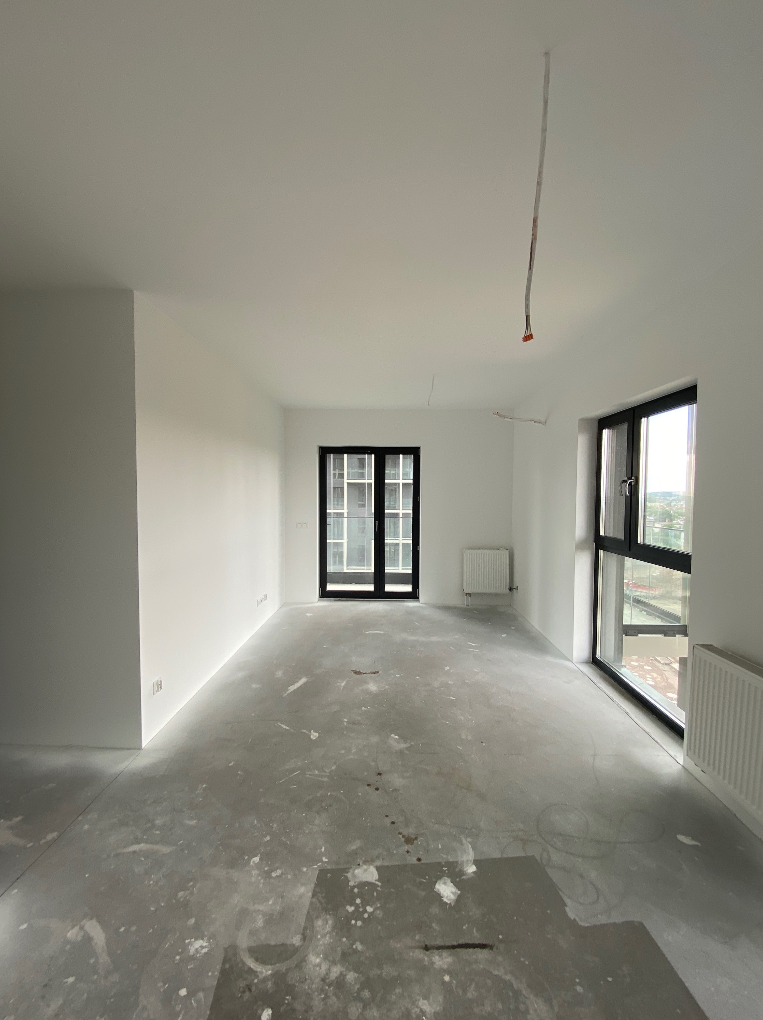 Pusty pokój z białymi ścianami po malowaniu, widoczne ślady farby na podłodze, grzejniki i okna z czarnymi ramami.