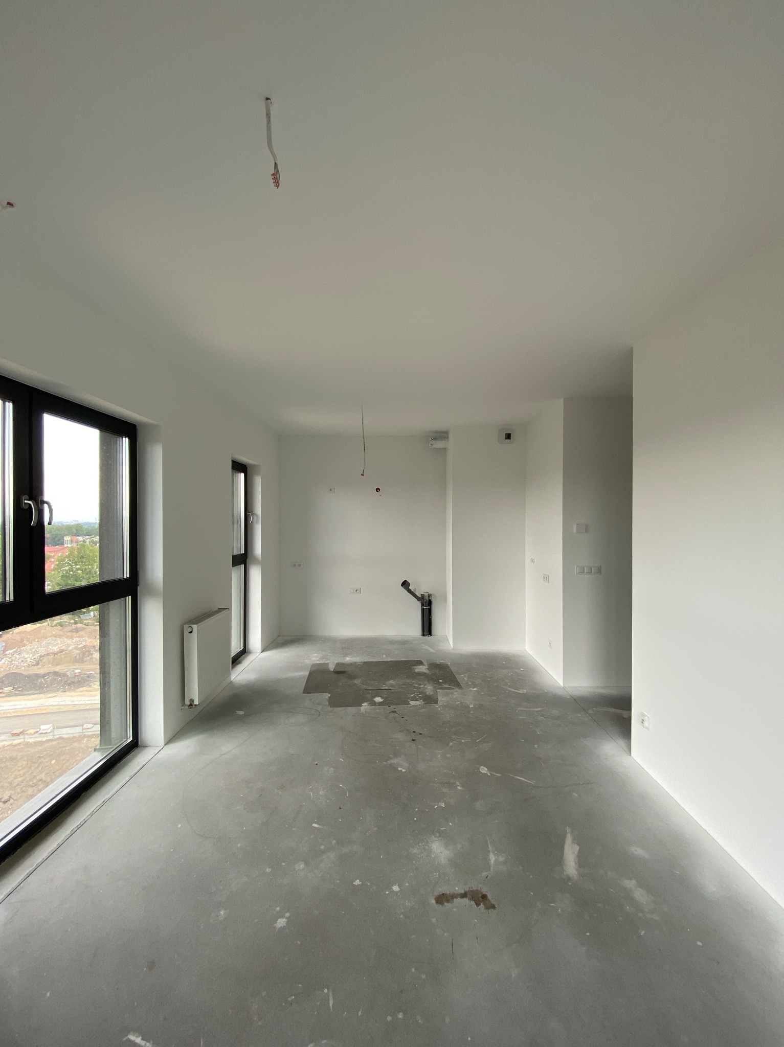 Surowe wnętrze nowego mieszkania z surowym betonem na podłodze, świeżo pomalowanymi na biało ścianami, dużymi oknami z czarnymi ramami i wystającymi kablami ze ściany i sufitu.