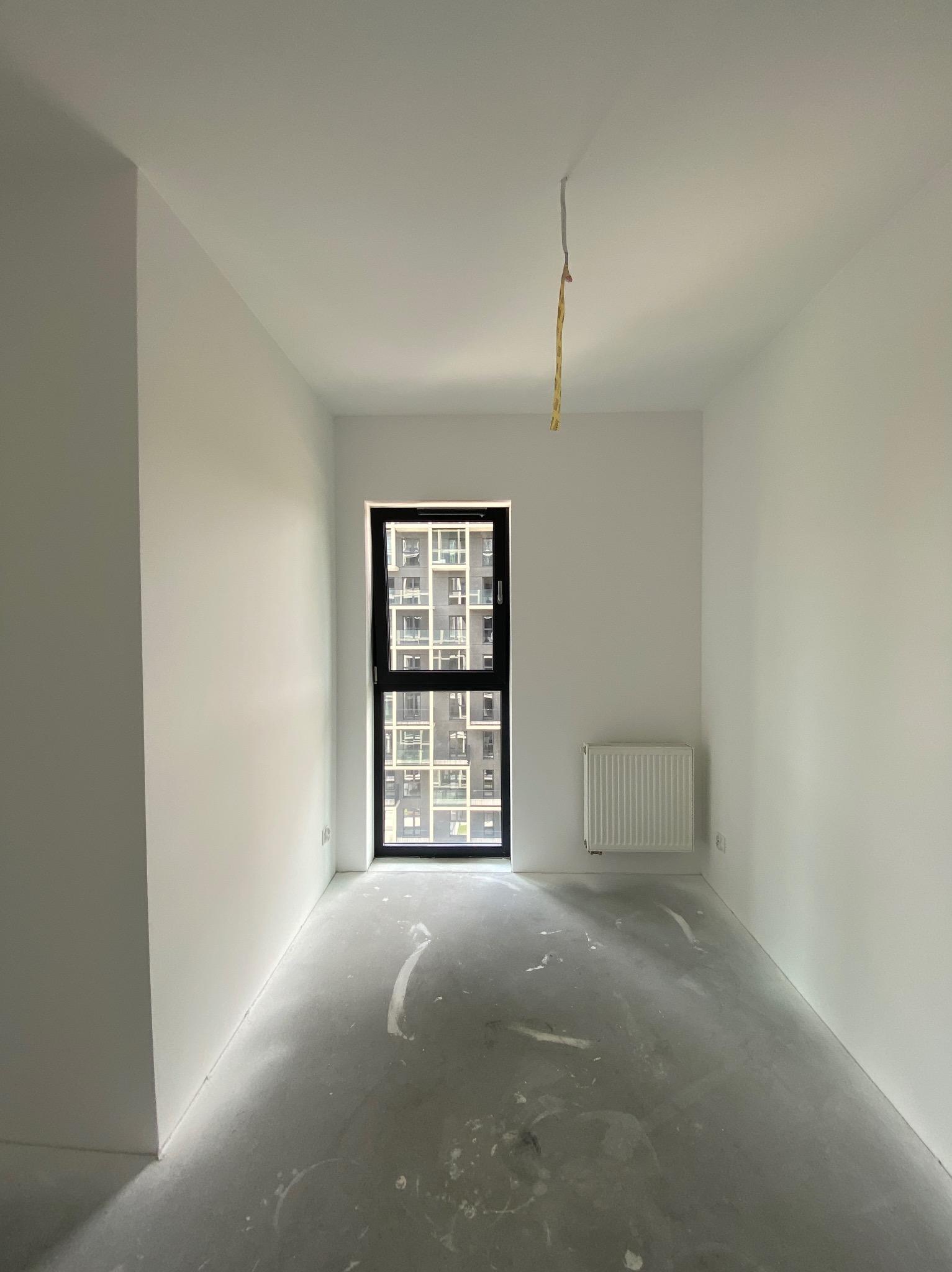 Pusty pokój w stanie deweloperskim z grzejnikiem, oknem z widokiem na budynek i wystającymi przewodami z sufitu.