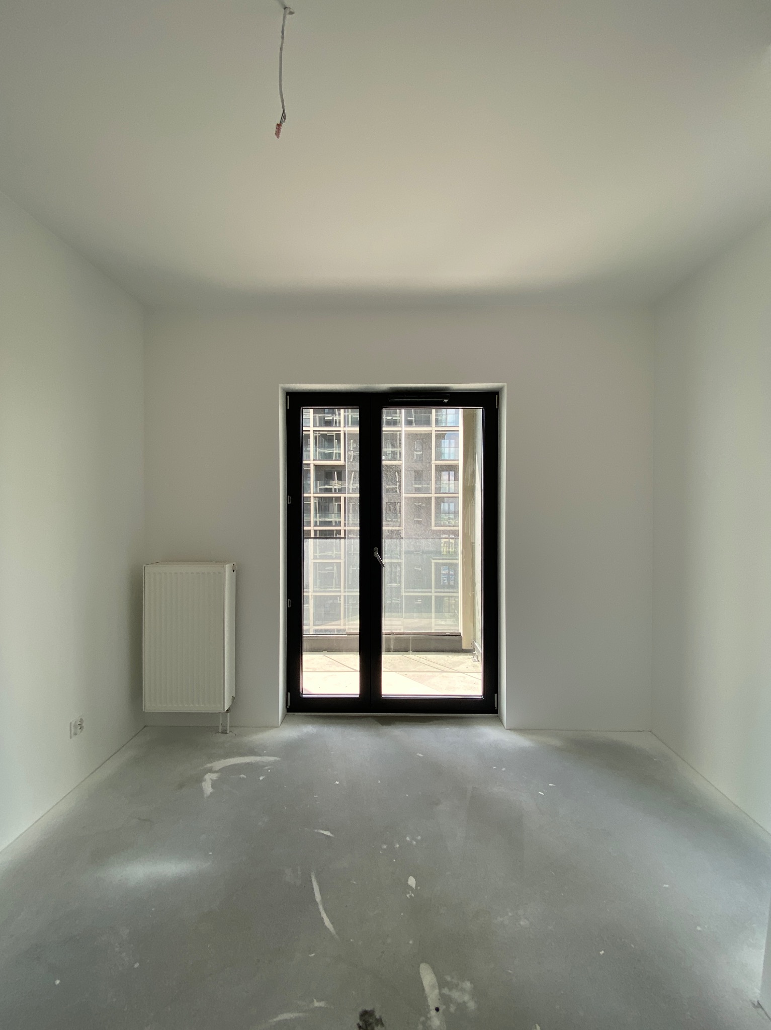 Pusty, jasny pokój z białymi ścianami, grzejnikiem i betonową podłogą w trakcie prac przygotowawczych, widok na budynek przez balkonowe drzwi.
