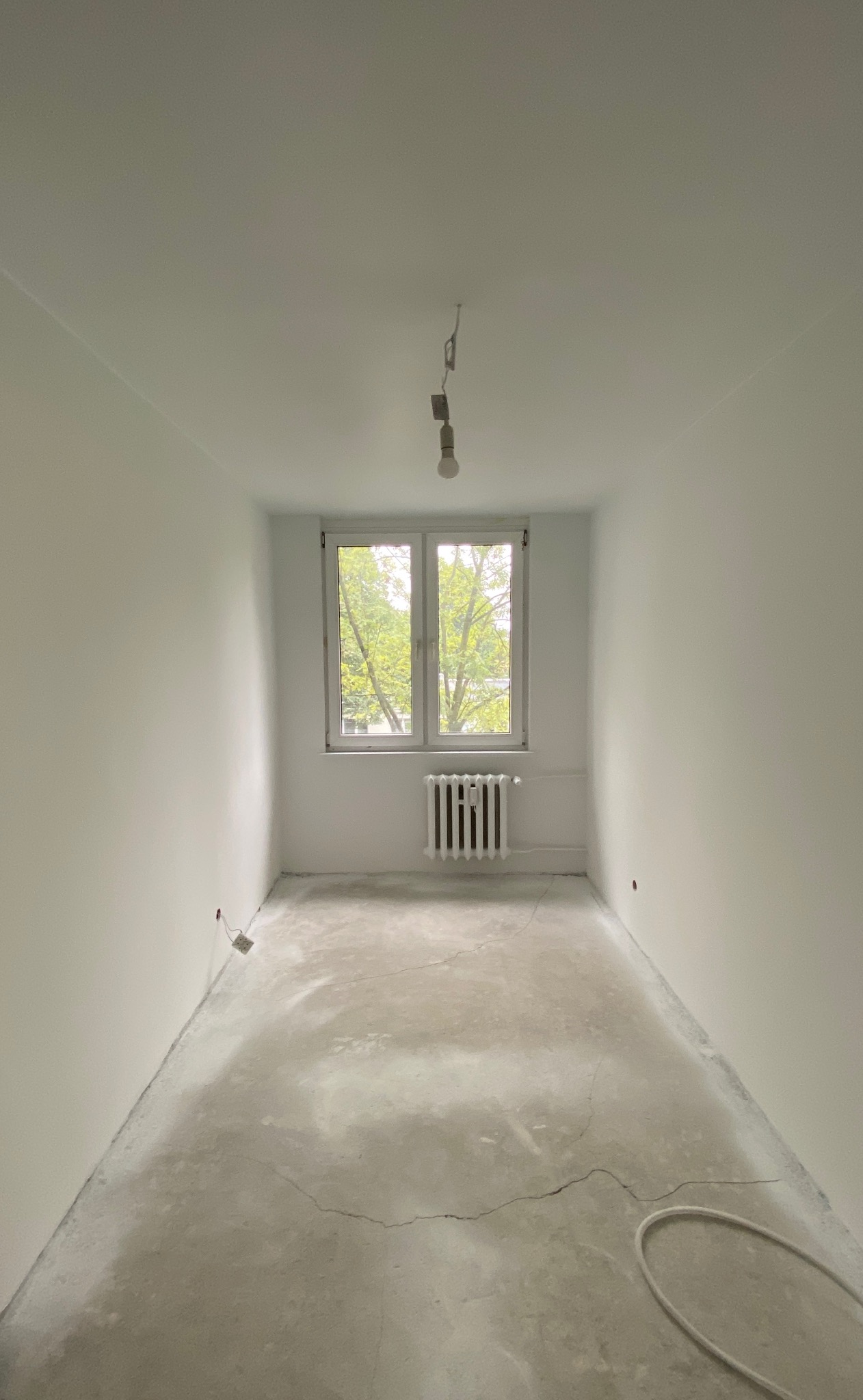 Pusty pokój po świeżym malowaniu na biało, widoczna surowa podłoga z pęknięciami, grzejnik pod oknem i wisząca żarówka.