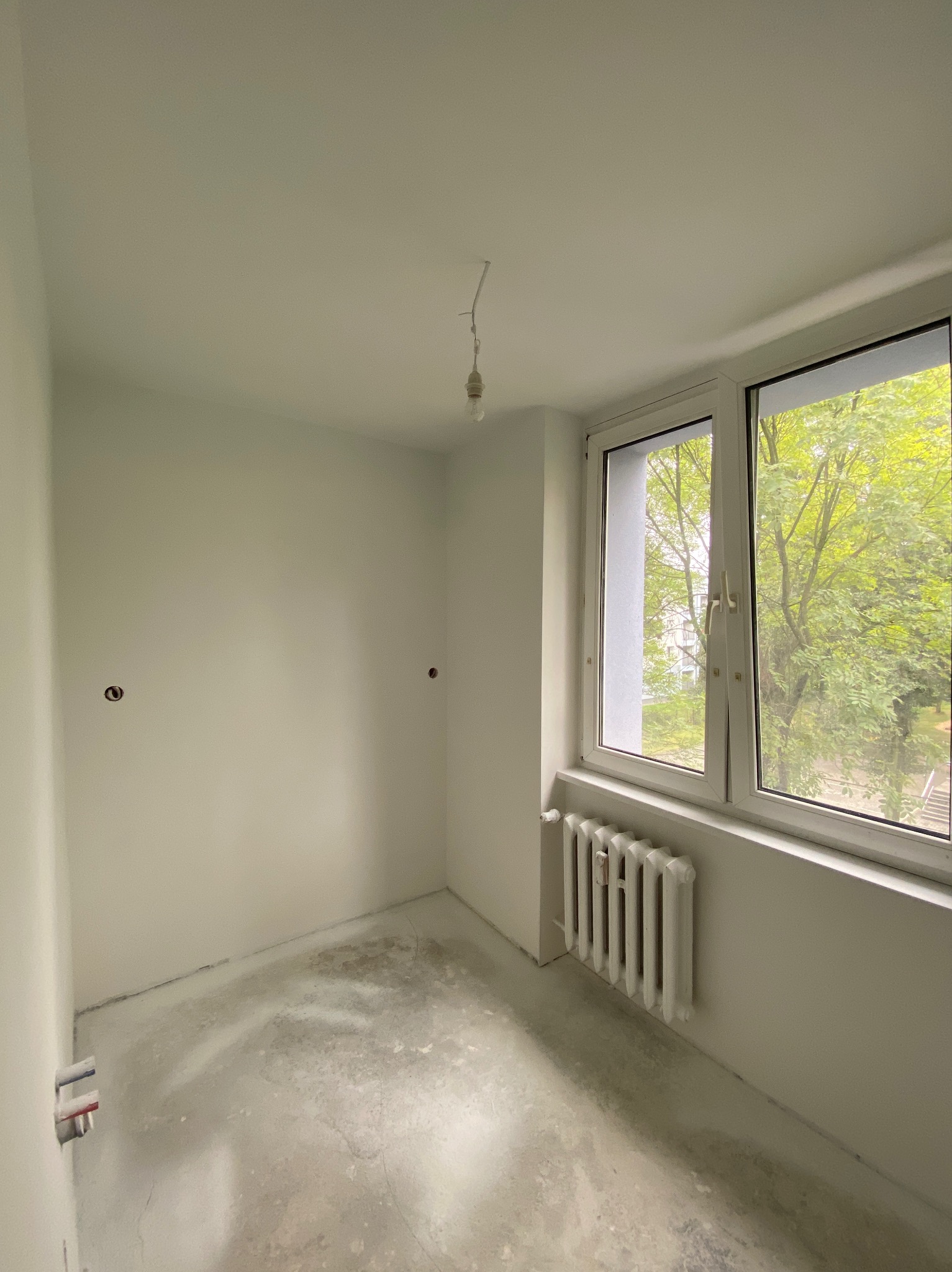 Pusty, odświeżony pokój z białymi ścianami, betonową podłogą przygotowaną do dalszych prac, grzejnikiem pod oknem i wystającymi kablami elektrycznymi.