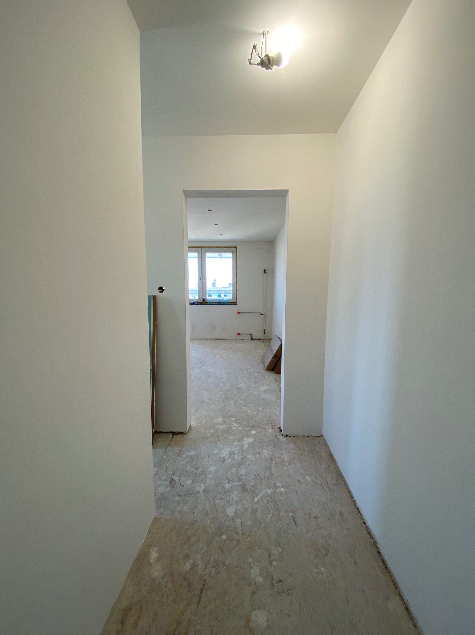 Korytarz w mieszkaniu po świeżym pomalowaniu ścian na biało, widoczne surowe podłoże podłogi i przejście do drugiego pokoju z oknem.