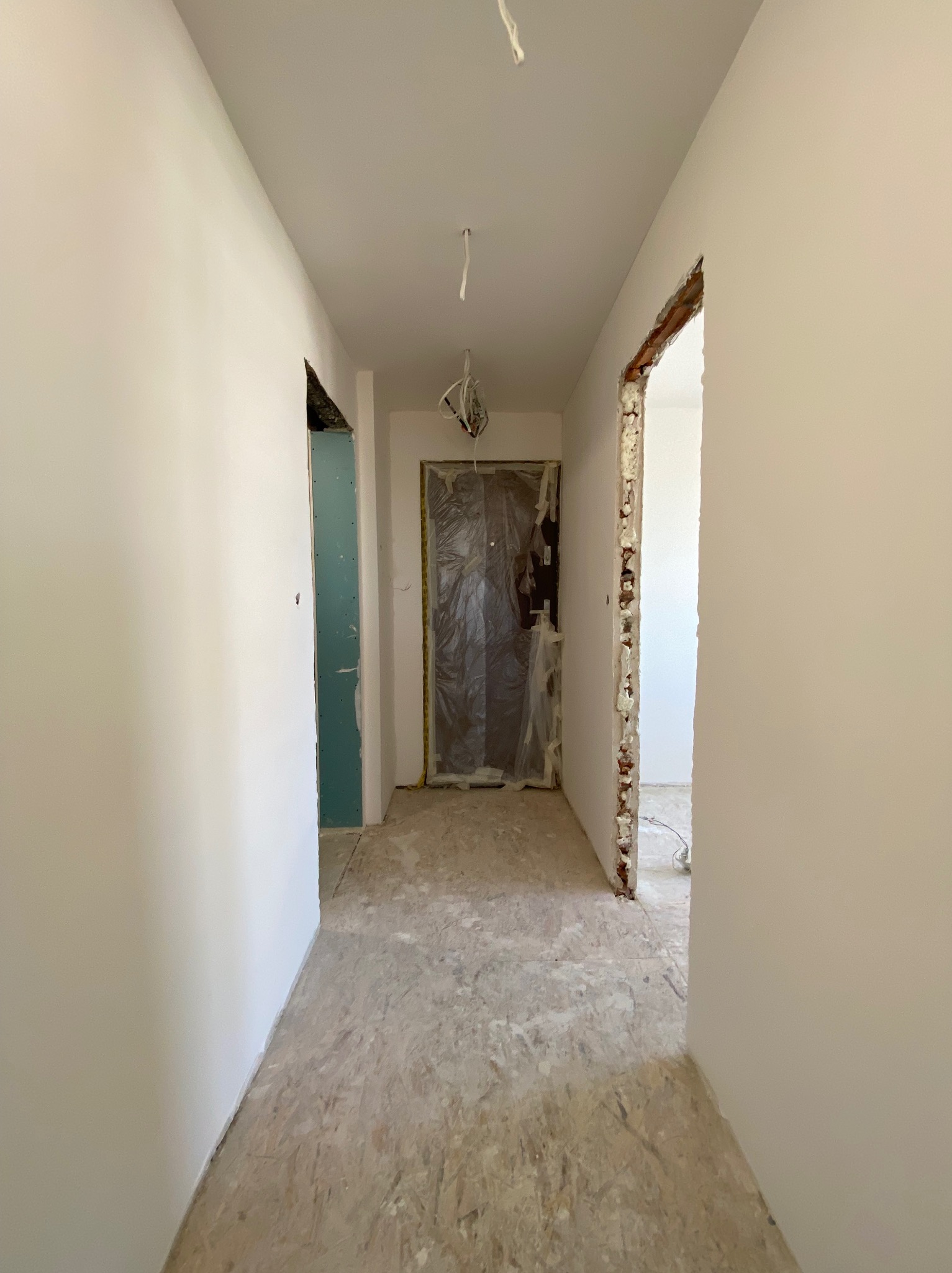 Korytarz w trakcie prac: ściany po szpachlowaniu i malowaniu na biało, widoczne fragmenty płyty karton gips, zabezpieczone folią drzwi.