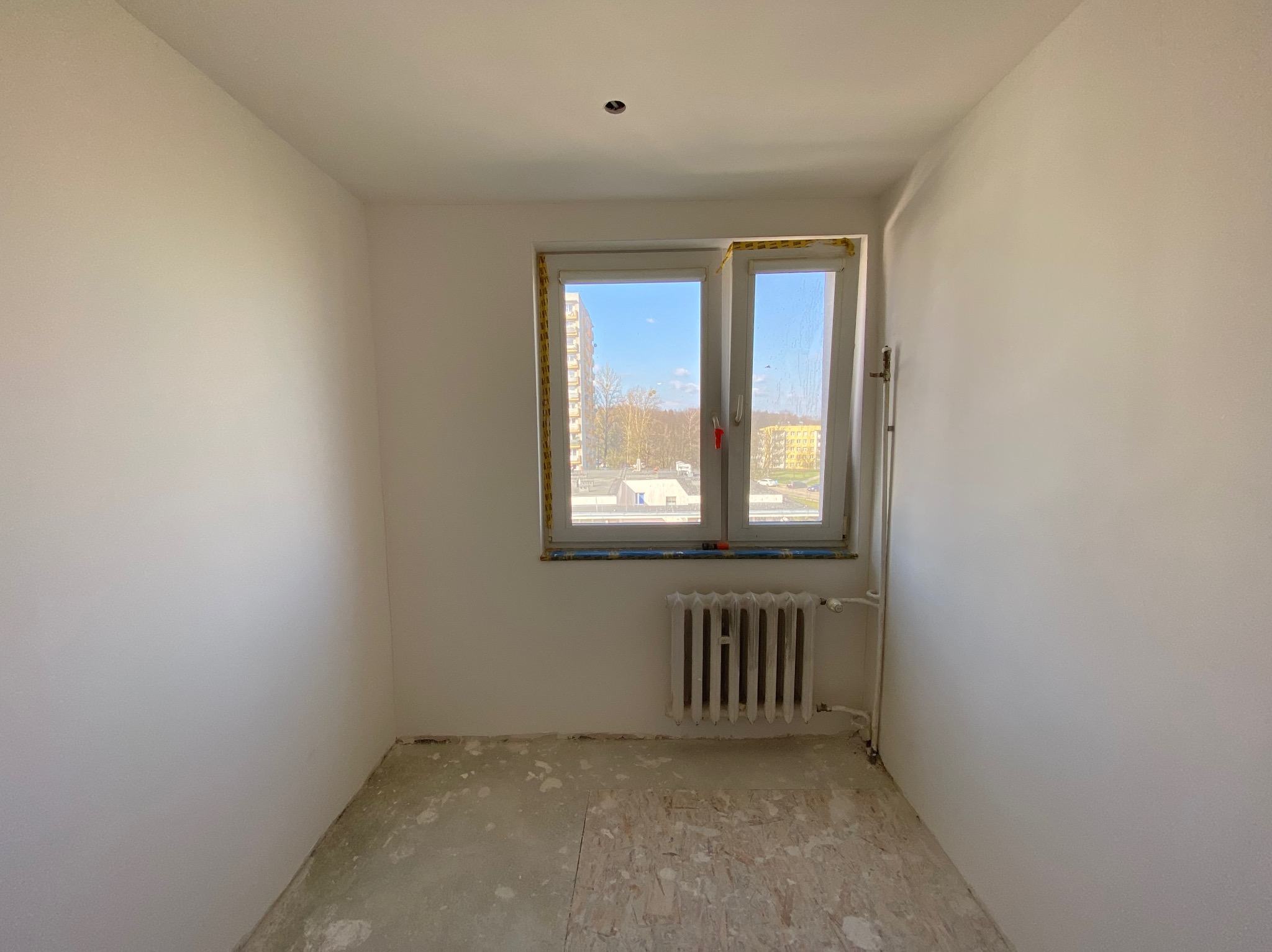 Pusty pokój przed pracami wykończeniowymi, z oknem zabezpieczonym taśmą malarską i starym grzejnikiem, ślady po poprzednich warstwach na podłodze.