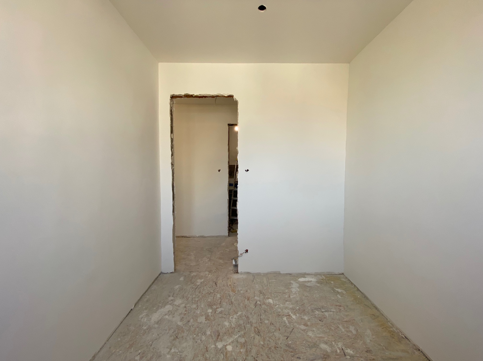 Wnętrze pokoju po szpachlowaniu ścian, widoczne gładkie, białe powierzchnie i niezabezpieczona podłoga z płyty OSB oraz otwór drzwiowy w stanie surowym.