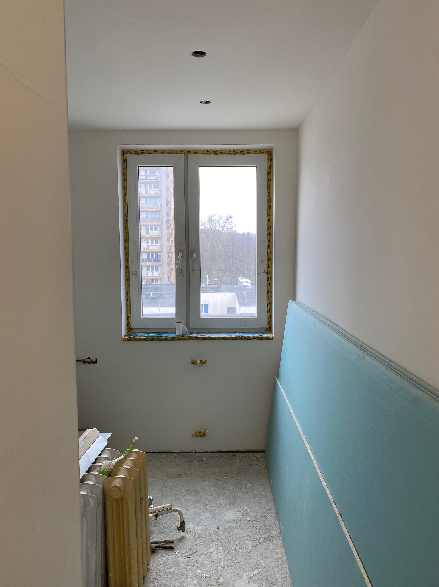 Wnętrze pokoju w trakcie remontu: ściany przygotowane do malowania, płyty karton-gips o niebieskim zabarwieniu oparte o ścianę, okno zabezpieczone taśmą malarską, widoczne przygotowane punkty...