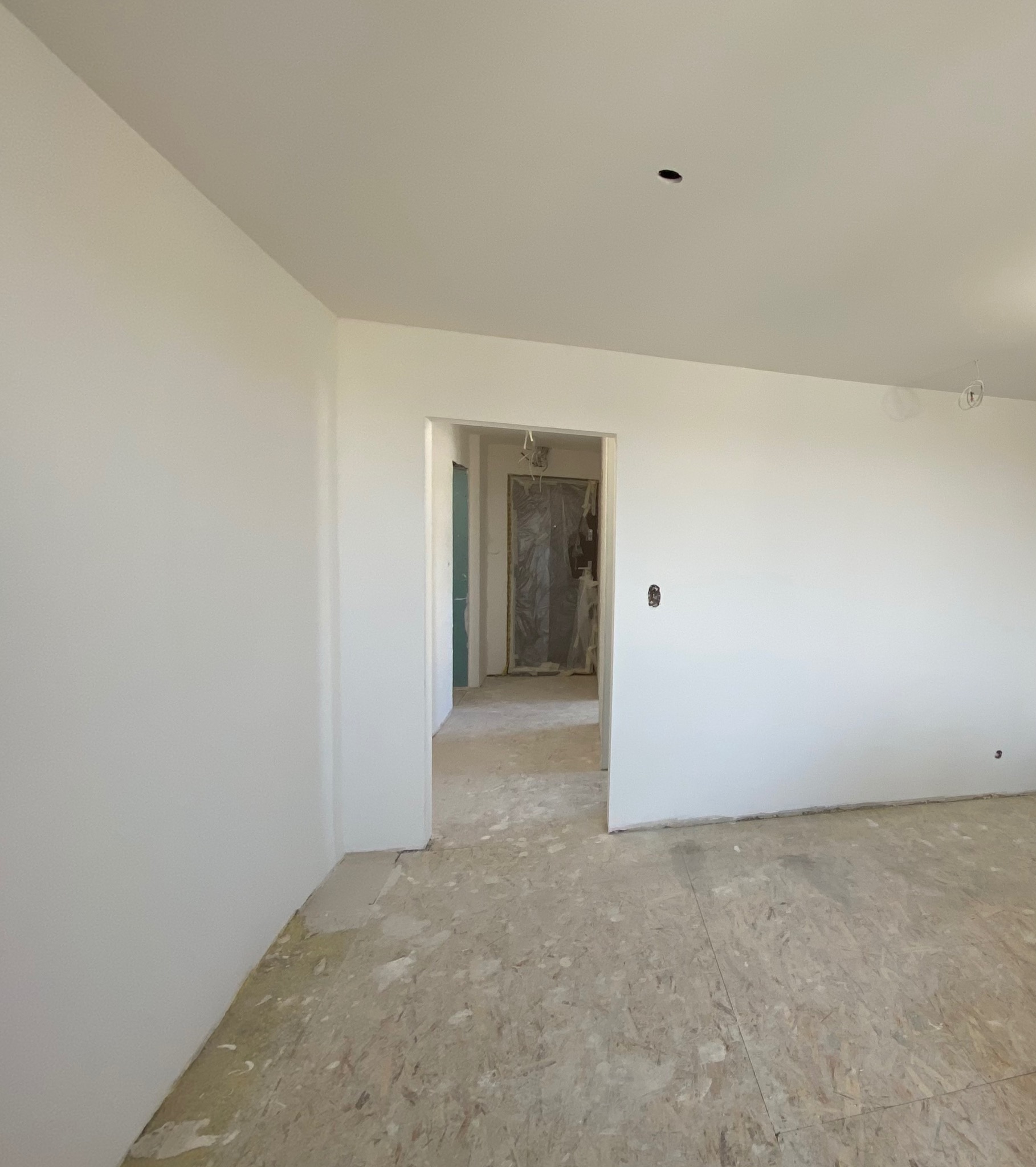 Wnętrze pokoju w trakcie prac przygotowawczych: świeżo pomalowane ściany na biało, surowa podłoga, widoczne pozostałości po pracach budowlanych, zabezpieczone folią drzwi w tle.