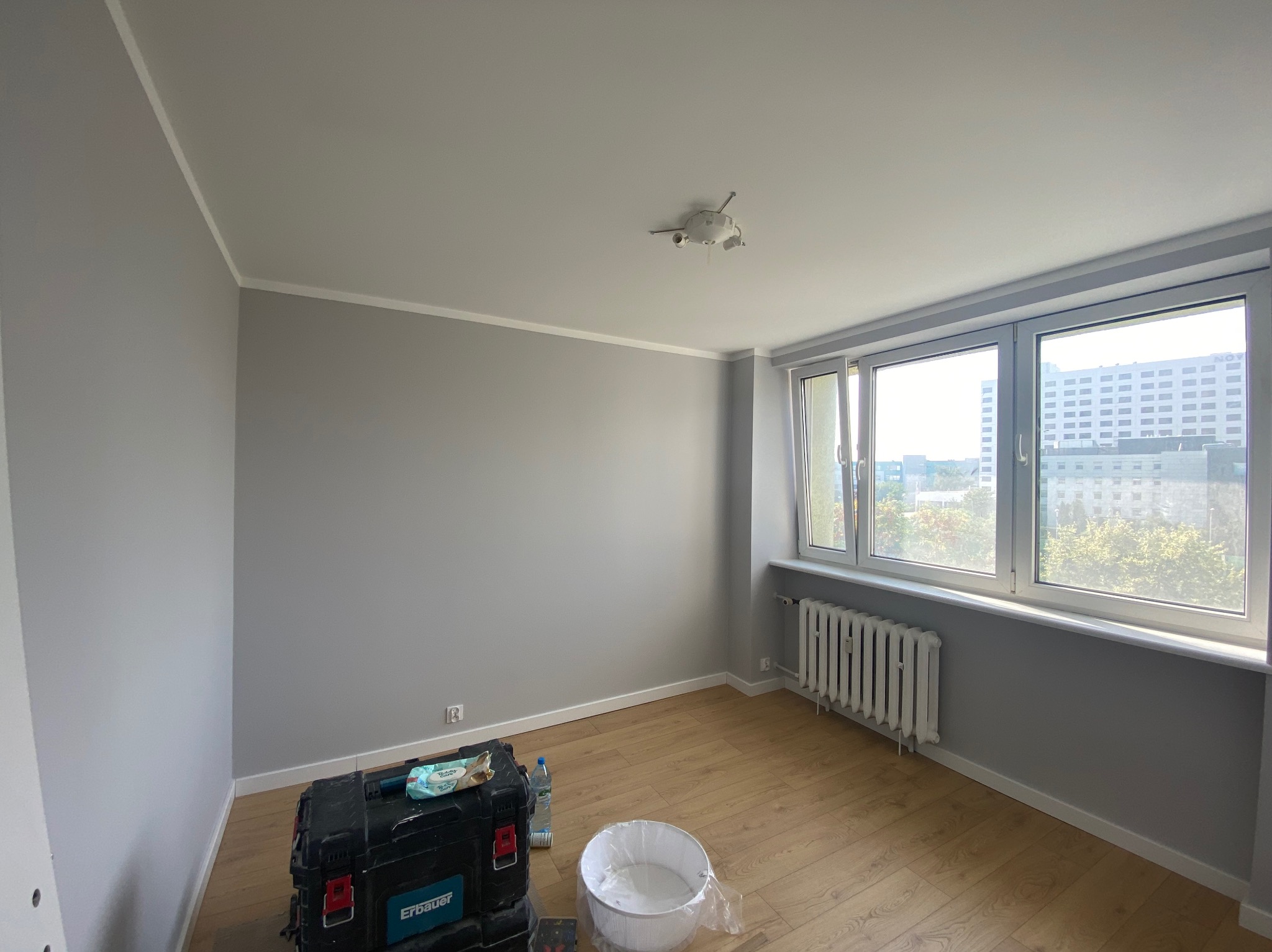 Pomieszczenie po malowaniu ścian na szaro, widoczny grzejnik, okno z widokiem na budynek i narzędzia malarskie na podłodze.