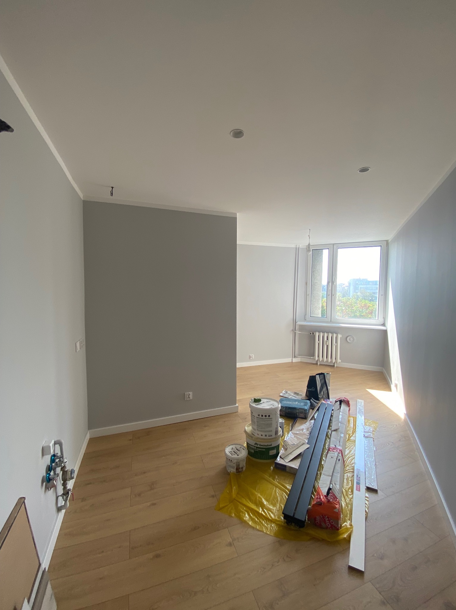 Wnętrze mieszkania w trakcie odświeżania: świeżo pomalowane szare ściany, drewnopodobna podłoga, materiały budowlane rozłożone na folii ochronnej, widoczne puszki z farbą, listwy przypodłogowe...