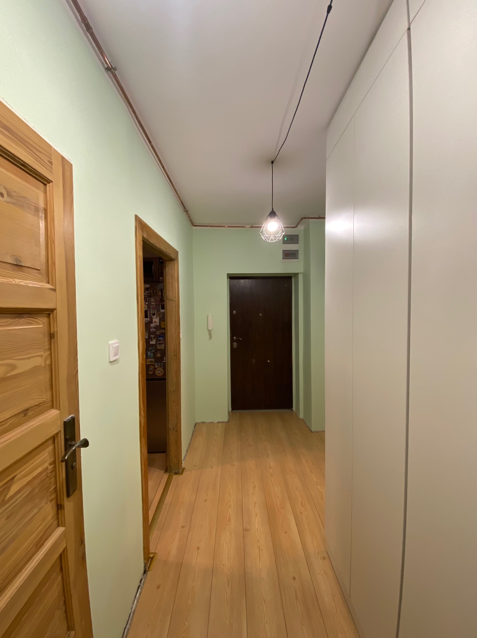 Korytarz po odświeżeniu: ściany pomalowane na jasnozielono, drewniana podłoga i drzwi, minimalistyczna lampa wisząca, zabudowa szafy wnękowej.