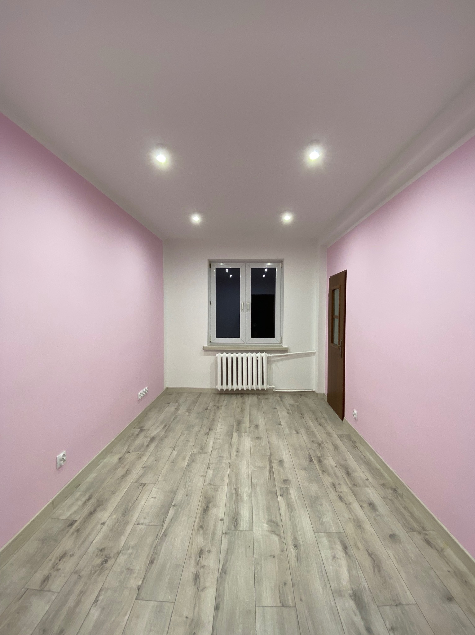 Pomalowane na różowo wnętrze pokoju z panelami podłogowymi, białym oknem i grzejnikiem oraz brązowymi drzwiami.