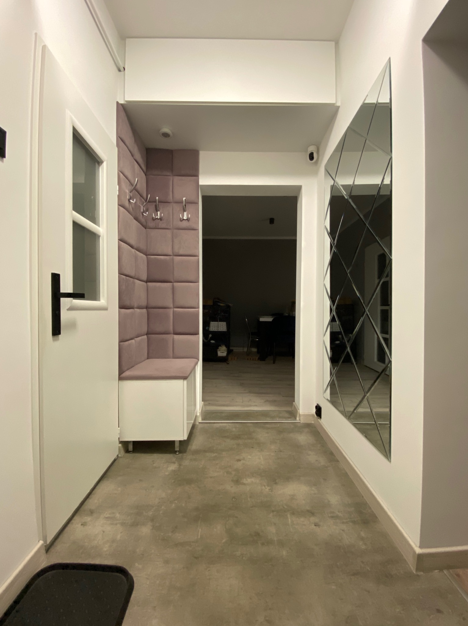 Przedpokój z siedziskiem obitym fioletową tapicerką w kwadraty, wieszakami i lustrem złożonym z rombów, widok na dalszą część mieszkania.