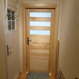 Zamknięte, jasne, drewniane drzwi wewnętrzne z trzema poziomymi przeszkleniami, widoczne w korytarzu z szarą wycieraczką i drugimi białymi drzwiami po lewej stronie.