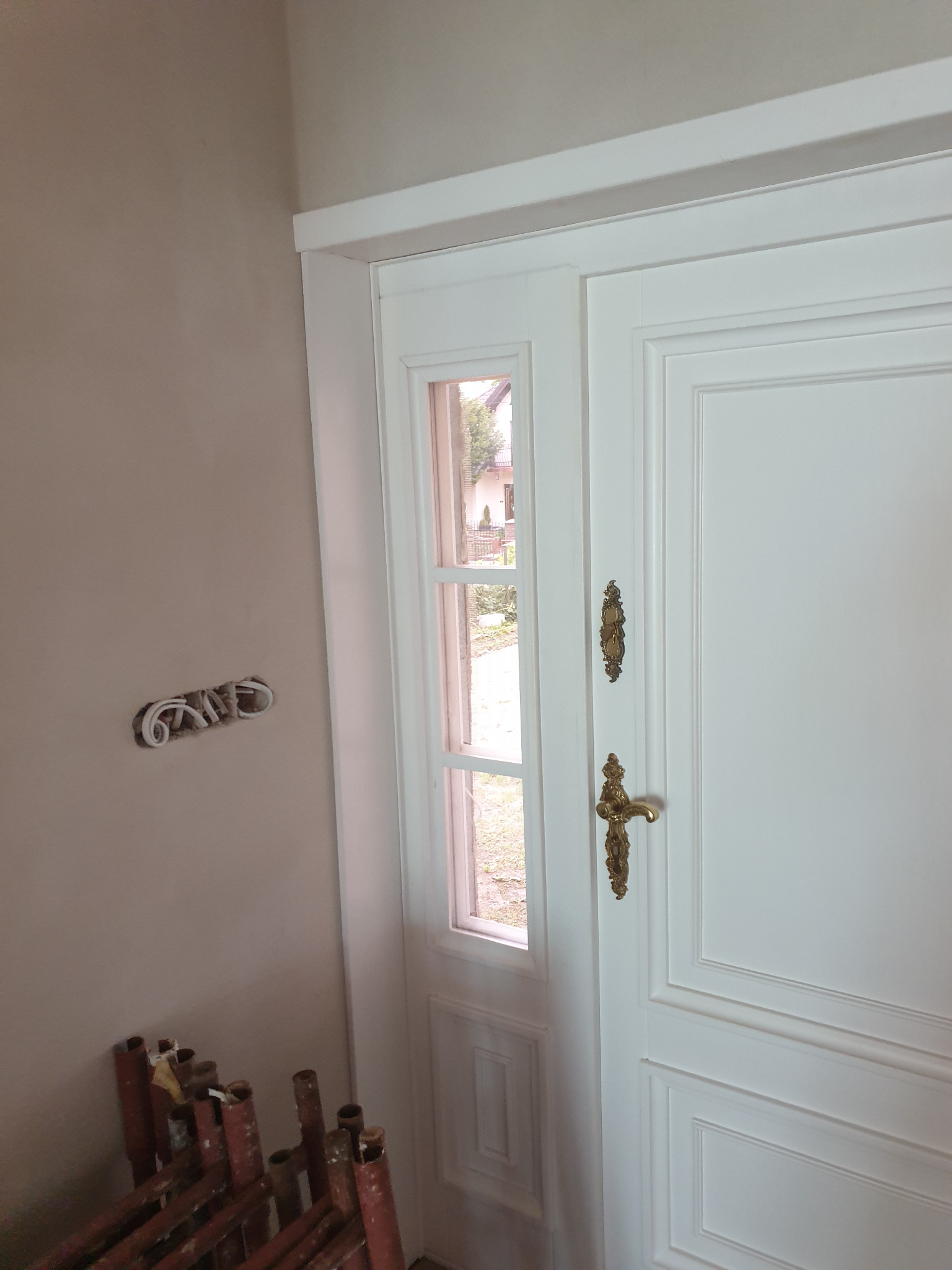 Białe, panelowe drzwi wejściowe z małym okienkiem, zdobione złotymi okuciami, widoczne na tle ściany z wystającymi przewodami elektrycznymi i fragmentem rusztowania.