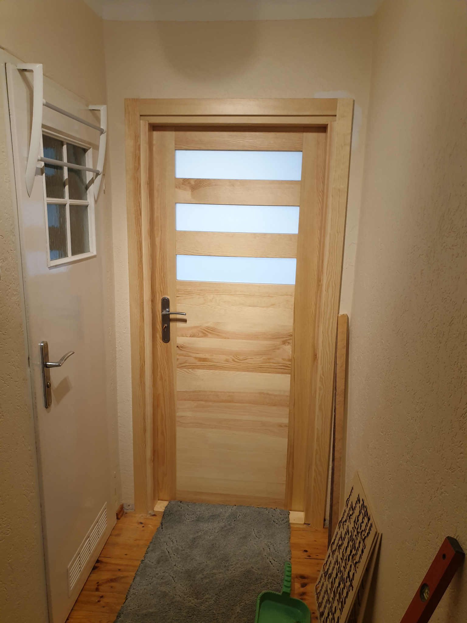 Zamknięte, jasne, drewniane drzwi wewnętrzne z trzema poziomymi przeszkleniami, widoczne w korytarzu z szarą wycieraczką i drugimi białymi drzwiami po lewej stronie.