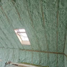 Poddasze z oknem dachowym, ocieplone pianką PUR w kolorze jasnozielonym, widoczne drewniane krokwie i elementy konstrukcyjne.