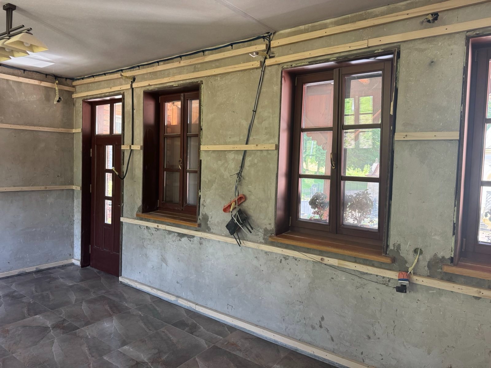 Przygotowanie ścian pod ocieplenie: surowe ściany z drewnianą konstrukcją, okna z brązowymi ramami, widoczne przewody i narzędzia. Wnętrze w trakcie prac.