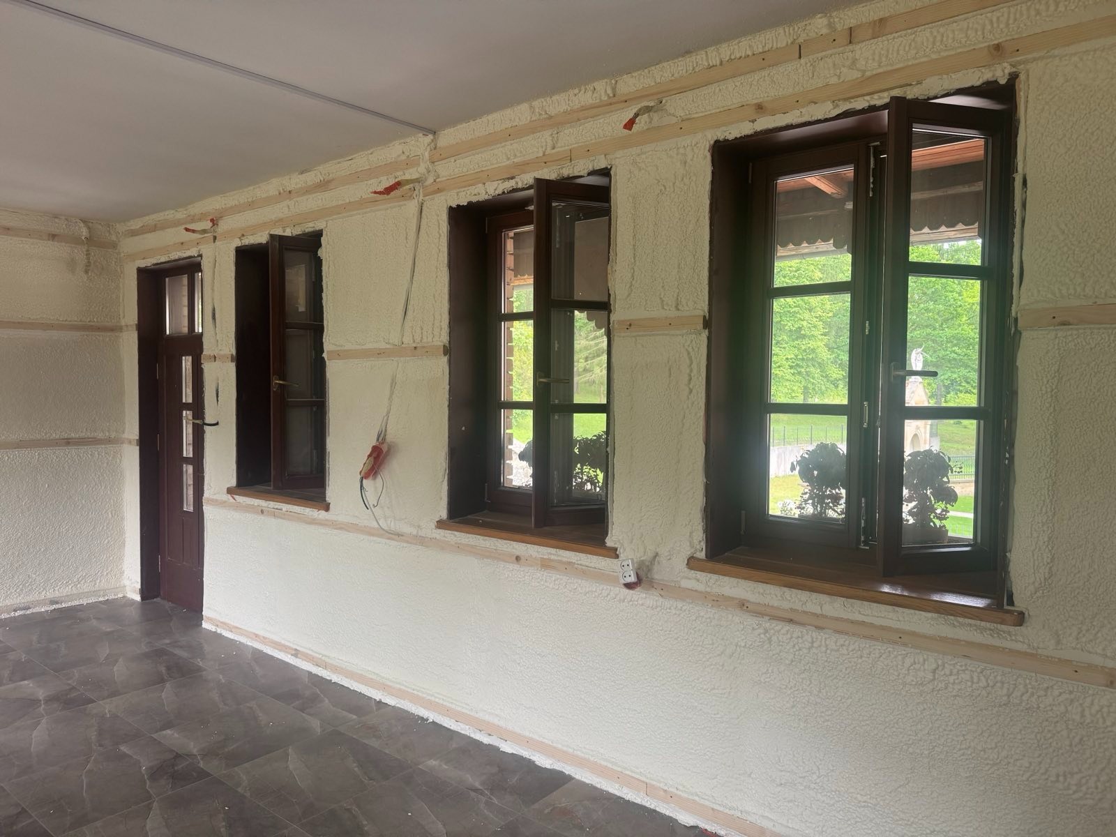 Poddasze po ociepleniu pianką PUR: widoczne drewniane ramy okienne z oknami, ściany i sufit pokryte białą pianką, listwy drewniane. Na ścianie gniazdko elektryczne.