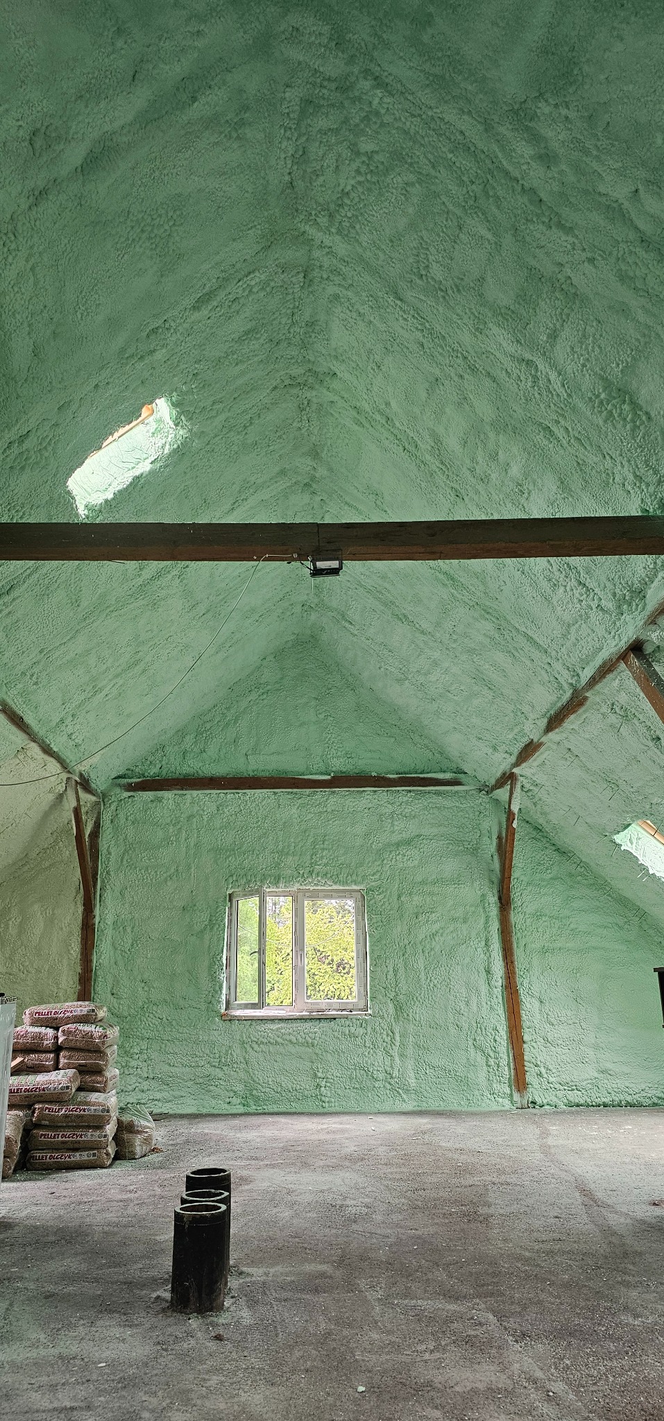 Poddasze po ociepleniu pianką PUR, widoczne drewniane elementy konstrukcyjne, okno i worki z pelletem. Izolacja w odcieniach zieleni pokrywa ściany i sufit.