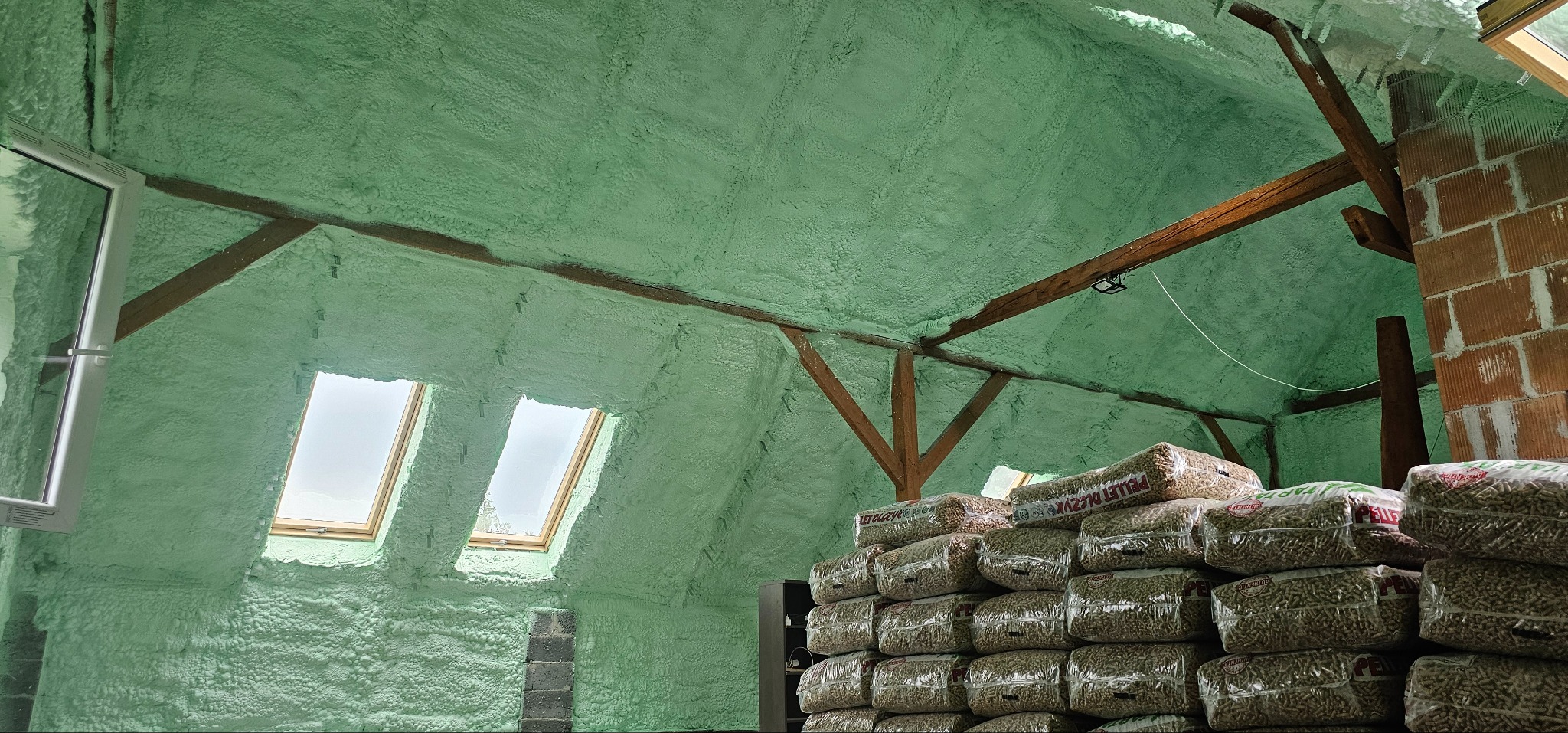 Poddasze po ociepleniu pianką PUR, widoczne okna dachowe i drewniana konstrukcja. Na pierwszym planie składowane worki z pelletem.