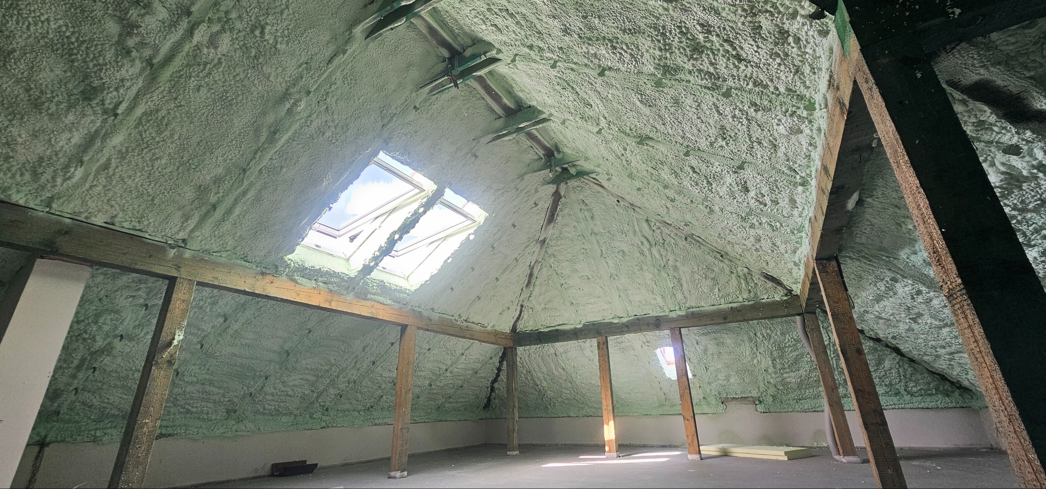 Poddasze po ociepleniu pianką PUR, widoczne drewniane elementy konstrukcyjne i okna dachowe. Jasna, równomierna warstwa izolacji pokrywa całą powierzchnię.