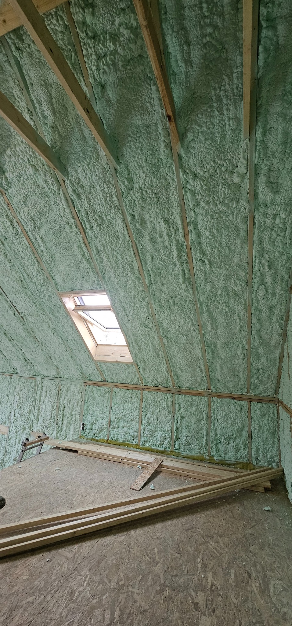 Poddasze z oknem dachowym, ocieplone pianką PUR w kolorze jasnozielonym, widoczne drewniane krokwie i elementy konstrukcyjne.