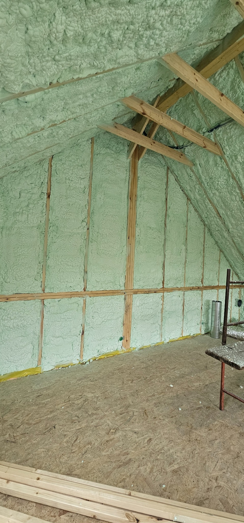 Poddasze z drewnianą konstrukcją i jasną pianką izolacyjną, widoczne fragmenty podłogi z płyty OSB i narzędzia. Ocieplenie wykonane z dbałością o detale.