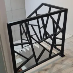 Nowoczesna balustrada schodowa w kolorze czarnym o geometrycznym wzorze, zamontowana w jasnym wnętrzu z drewnianą podłogą i białymi schodami.