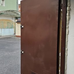 Krzysztof jarosz - Otwarta, brązowa brama garażowa z widocznym ociepleniem i uszczelnieniem pianką montażową, uchylona na zewnątrz budynku. Widoczne mocowania i zamek.