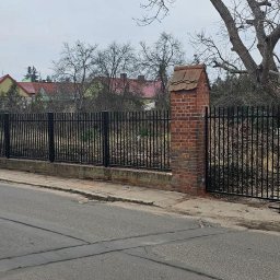 Krzysztof jarosz - Czarne, metalowe ogrodzenie posesji z bramą, wsparte na ceglanych słupkach z dachówką, widoczne z ulicy w pochmurny dzień.