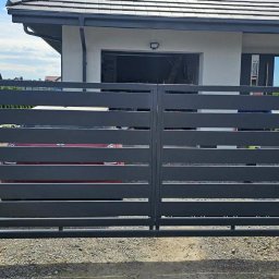Krzysztof jarosz - Nowoczesna, pozioma brama wjazdowa w kolorze grafitowym, zamontowana na posesji z szarym domem i kostką brukową. Widoczne betonowe słupki bramy.