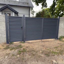 Krzysztof jarosz - Nowoczesna, grafitowa brama lamelowa z furtką, zamontowana między betonowymi słupkami imitującymi kamień. Widok na dom i zieleń w tle.
