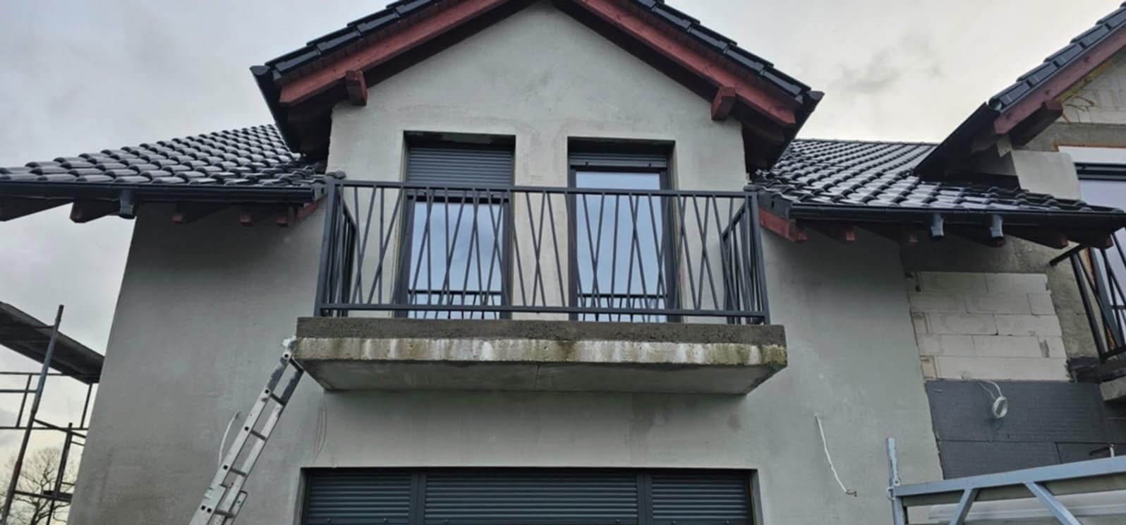 Balkon z czarną, metalową balustradą o geometrycznym wzorze, widoczny z dołu. Na balkonie dwa okna z roletami. Na ścianie domu widoczna drabina.