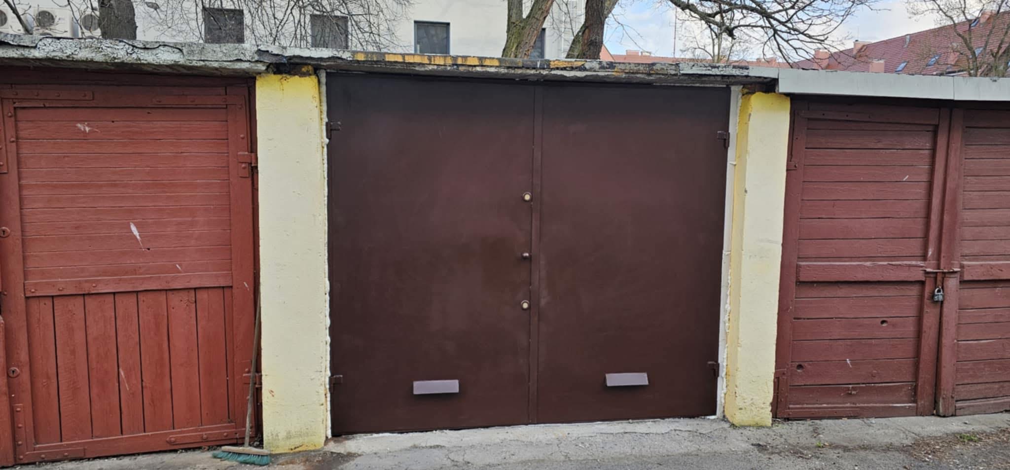 Zamykana, brązowa, dwuskrzydłowa brama garażowa, z widocznymi zamkami i uchwytami, umieszczona pomiędzy dwoma drewnianymi bramami w kolorze czerwonym.