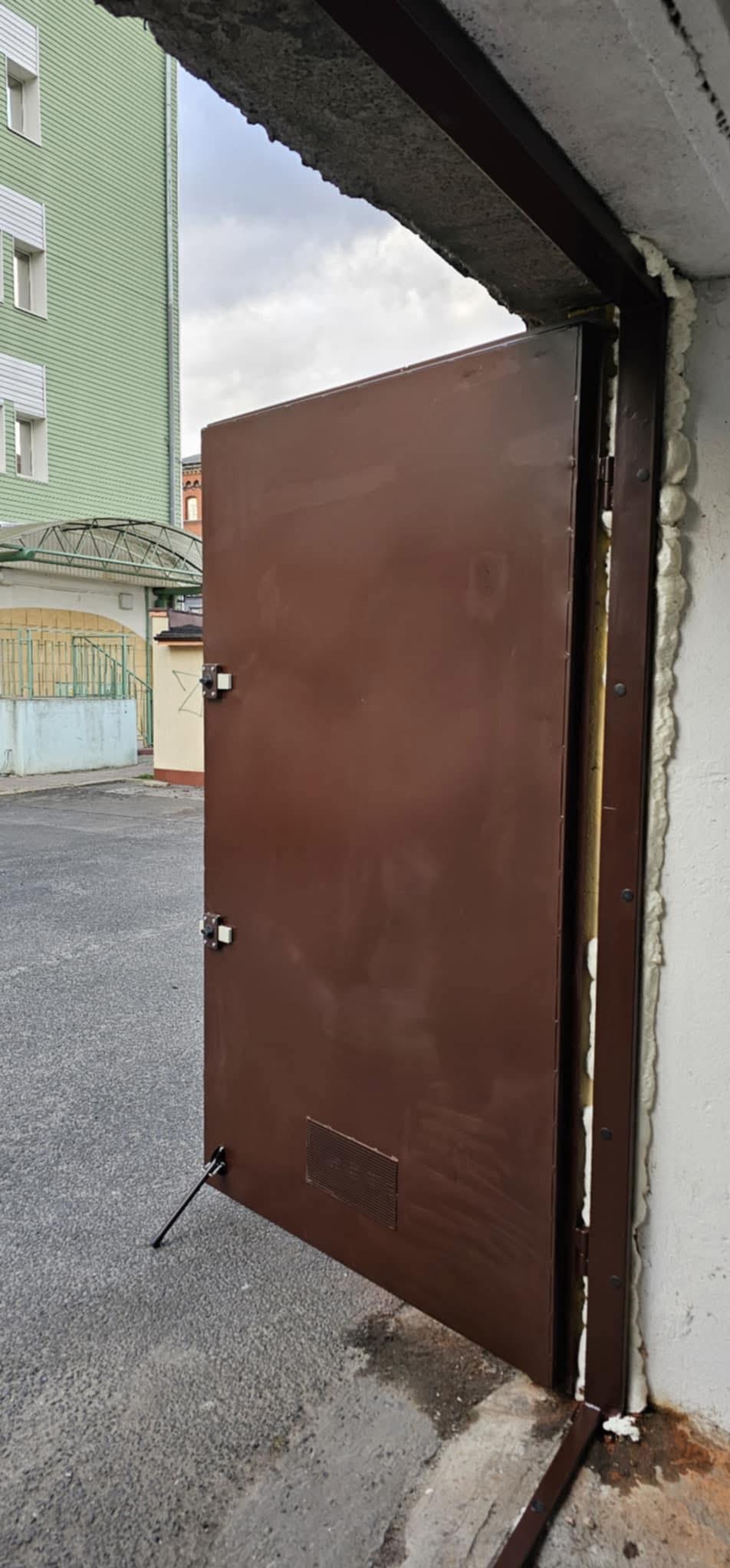 Otwarta, brązowa brama garażowa z widocznym ociepleniem i uszczelnieniem pianką montażową, uchylona na zewnątrz budynku. Widoczne mocowania i zamek.