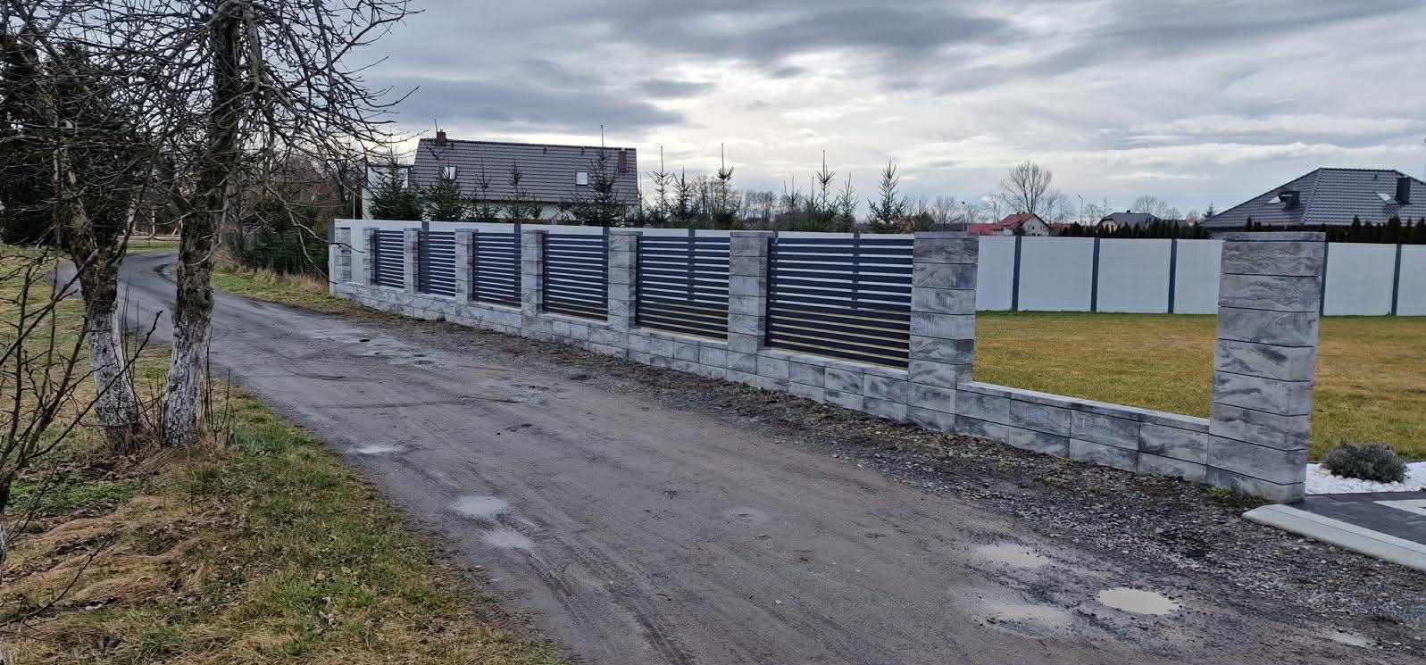 Nowoczesne ogrodzenie panelowe z szarego kamienia i poziomych, czarnych przęseł, widoczne z polnej drogi w pochmurny dzień. W tle dom z ciemnym dachem.