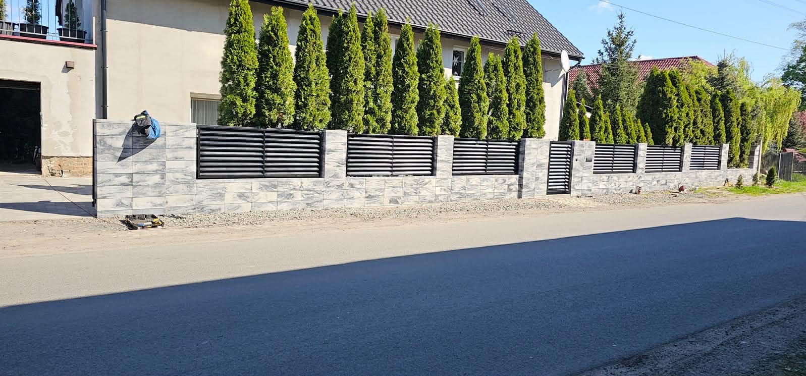 Nowoczesne ogrodzenie z szarej kostki i poziomych, czarnych paneli, z bramką wejściową. W tle rząd wysokich, zielonych tuj.