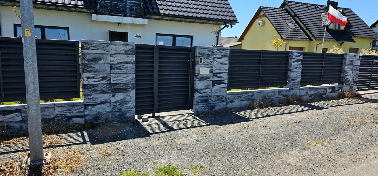 Nowoczesne ogrodzenie z poziomych, grafitowych paneli i kamiennych słupków, z bramą wjazdową na tle domów jednorodzinnych w słoneczny dzień. Widoczny fragment chodnika.