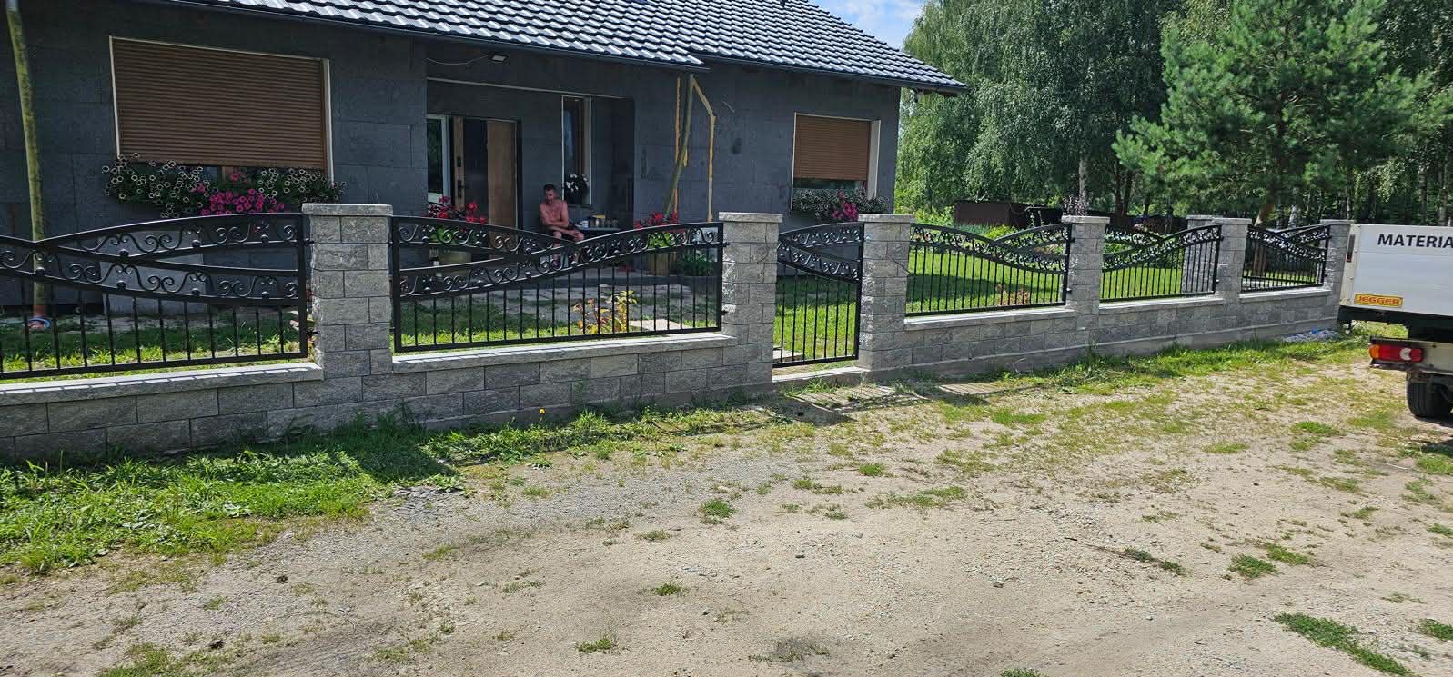 Czarno-szare ogrodzenie z kutymi elementami, otaczające posesję z szarym domem i zielonym ogrodem. Widoczny fragment samochodu dostawczego.