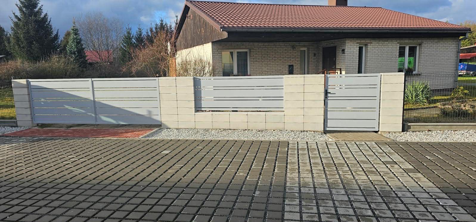 Nowoczesne ogrodzenie panelowe z bramą wjazdową, szare panele poziome, betonowe słupki, kostka brukowa na podjeździe, dom jednorodzinny w tle.