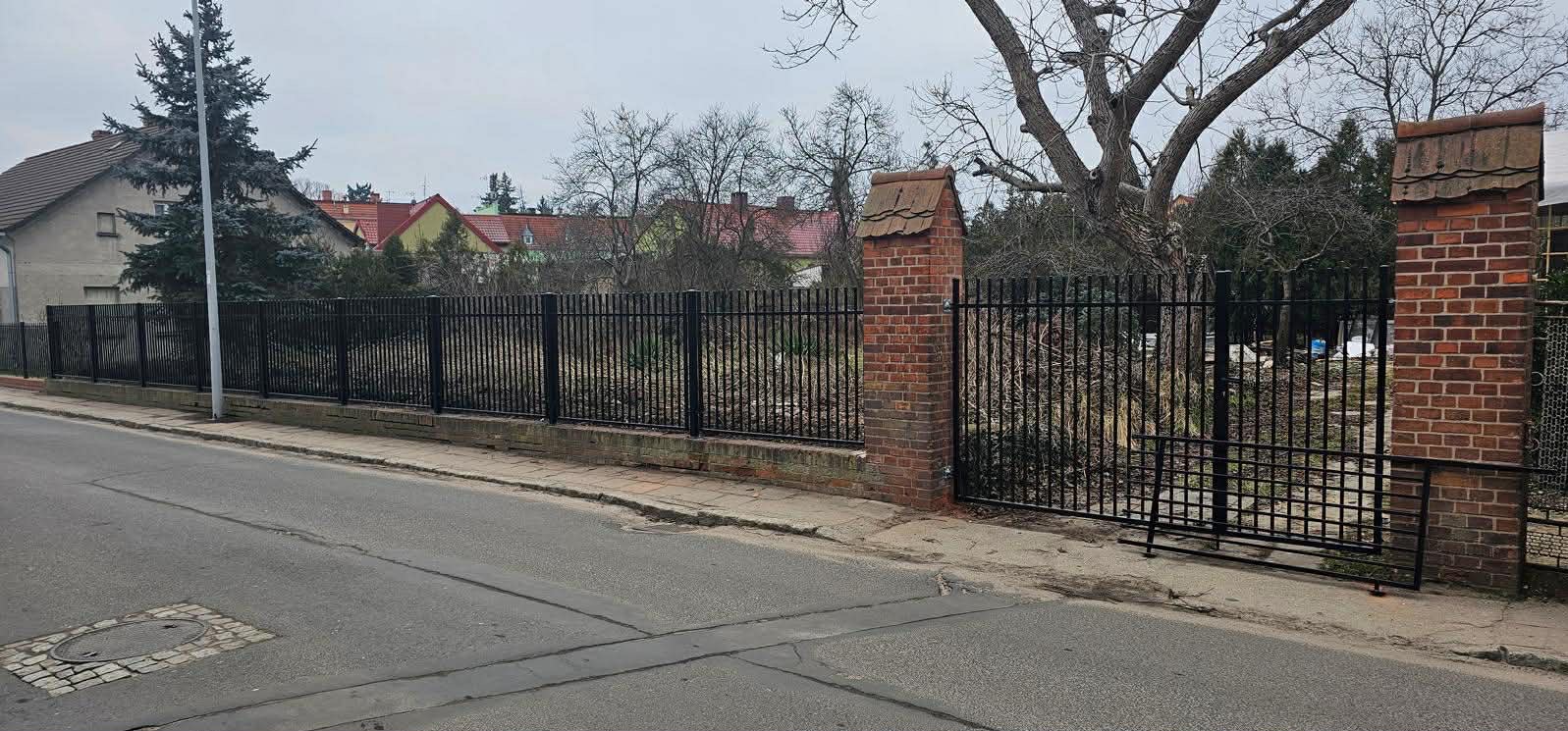 Czarne, metalowe ogrodzenie posesji z bramą, wsparte na ceglanych słupkach z dachówką, widoczne z ulicy w pochmurny dzień.