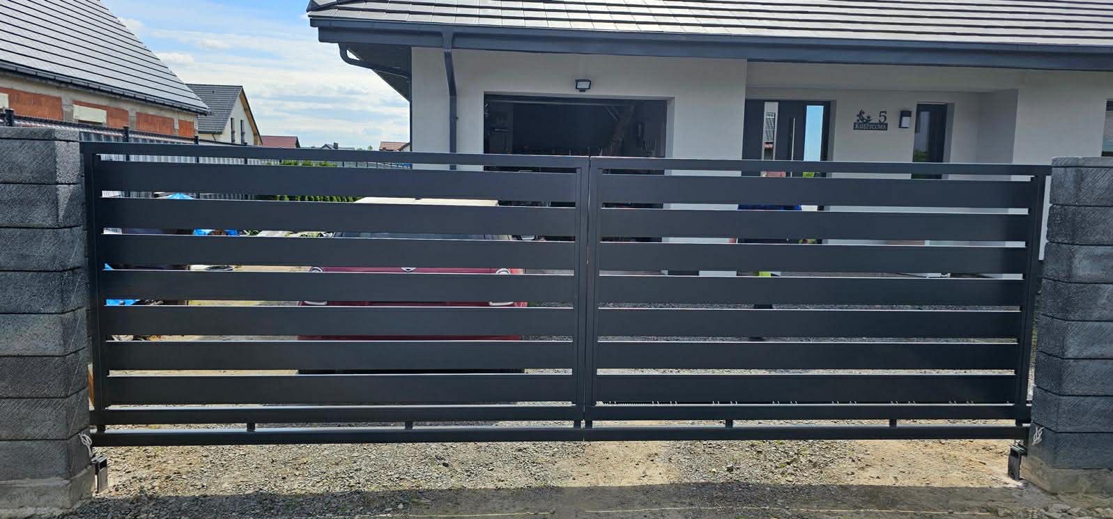 Nowoczesna, pozioma brama wjazdowa w kolorze grafitowym, zamontowana na posesji z szarym domem i kostką brukową. Widoczne betonowe słupki bramy.