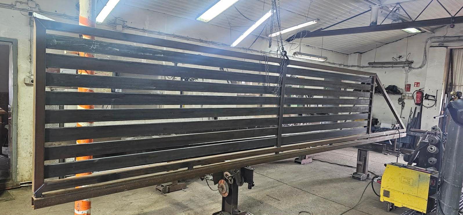 Surowa, pozioma brama wjazdowa z metalowych profili, w trakcie produkcji w warsztacie. Widoczne narzędzia spawalnicze i surowe wykończenie metalu.