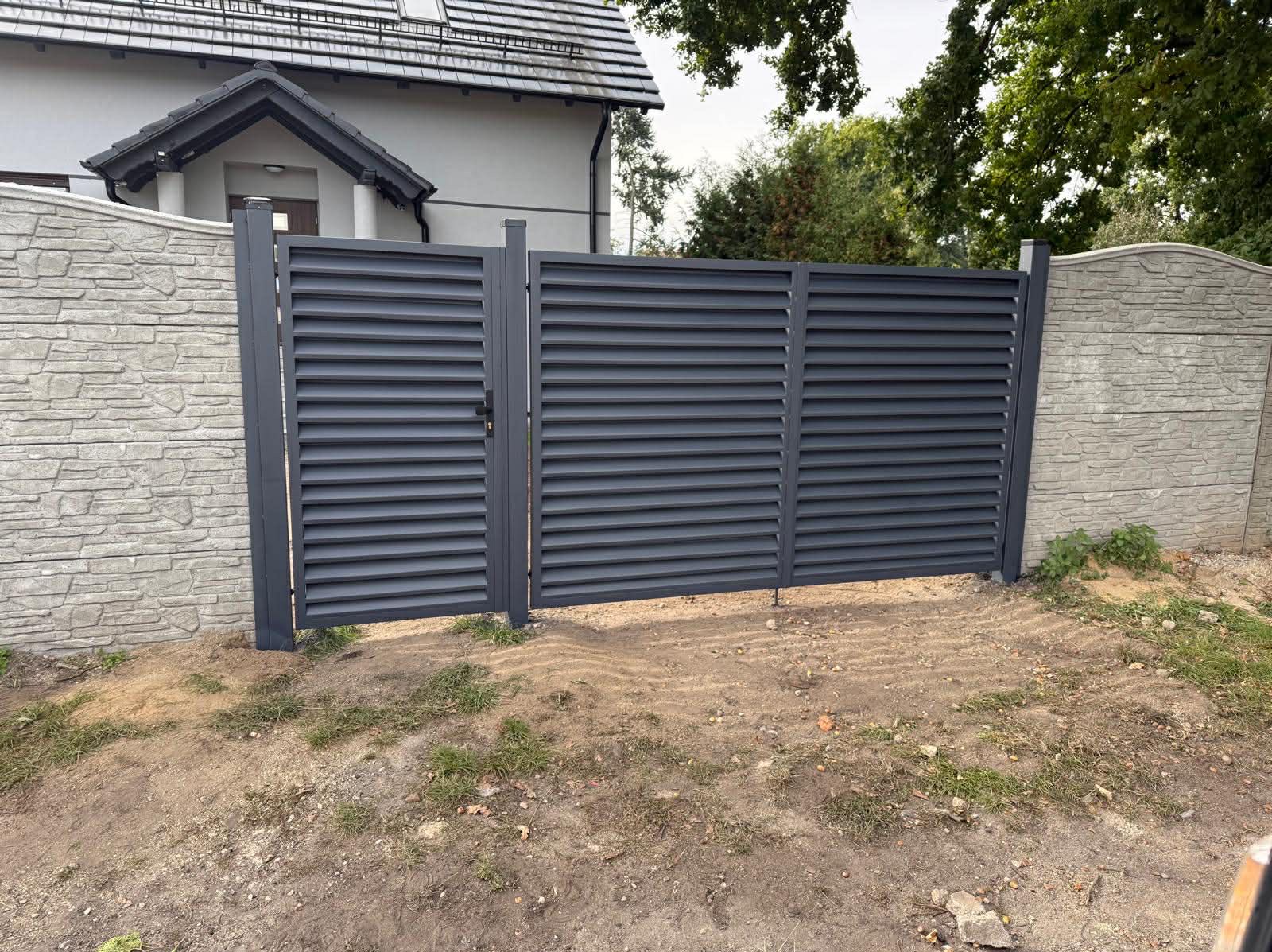 Nowoczesna, grafitowa brama lamelowa z furtką, zamontowana między betonowymi słupkami imitującymi kamień. Widok na dom i zieleń w tle.