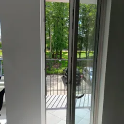 Otwarty balkon z widokiem na park. Białe płytki na podłodze balkonu i metalowa barierka. Za barierką widać zieleń parku i zaparkowane samochody.